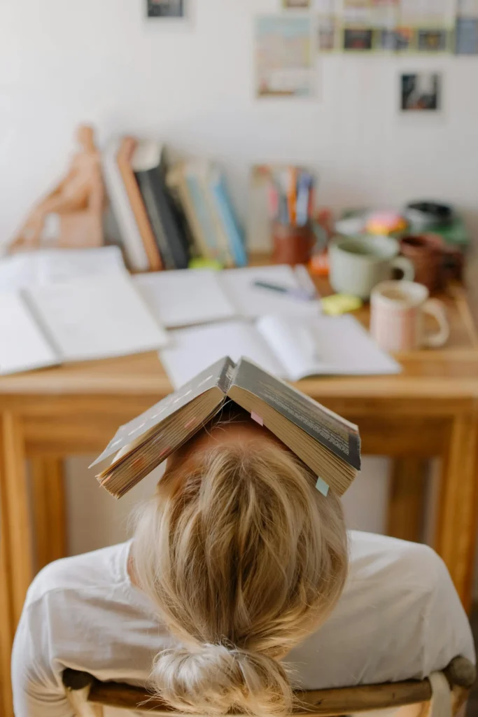 Jeune femme avec un livre ouvert sur sa tête, montrant un moment de fatigue ou de stress lié aux études.