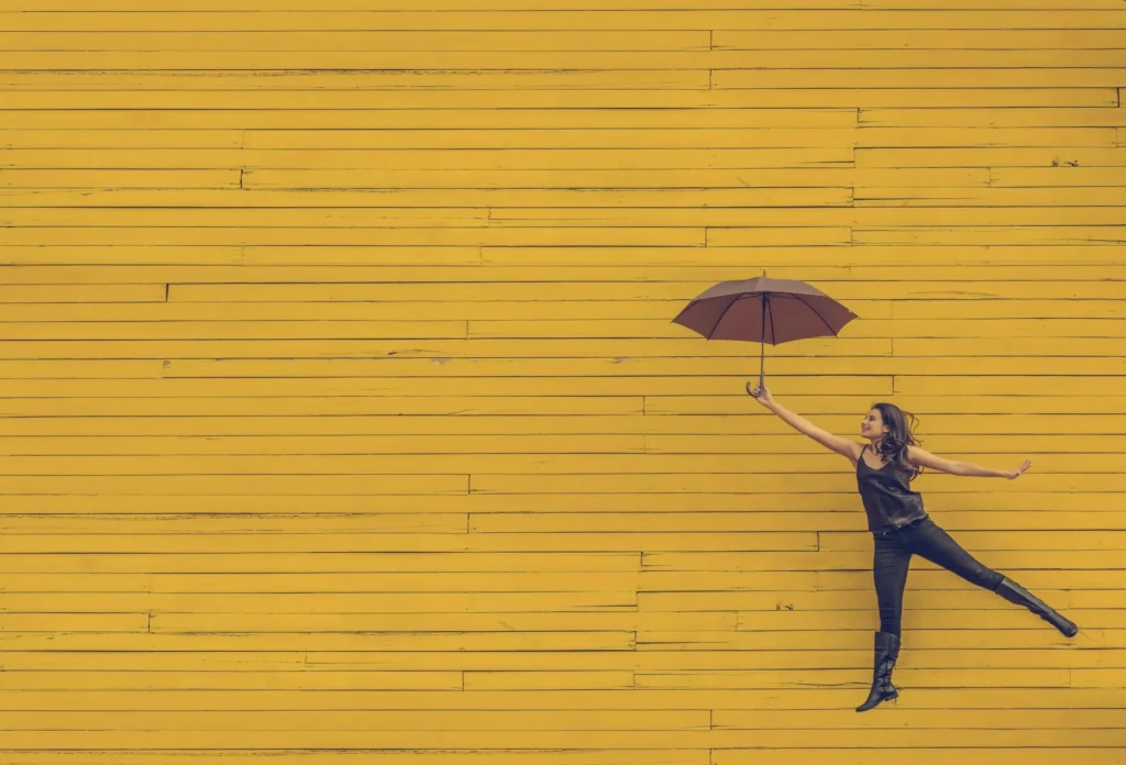 Jeune femme tenant un parapluie tout en dansant contre un mur jaune, illustrant la liberté, la créativité et le soutien dans le parcours d'accompagnement des jeunes.